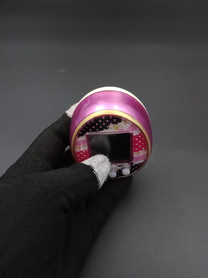 Puede incluir: Un juego electr&oacute;nico port&aacute;til rosa y blanco con una peque&ntilde;a pantalla y dos botones. El dispositivo tiene un borde decorativo con lunares y motivos florales. El dispositivo est&aacute; sostenido por una mano enguantada.