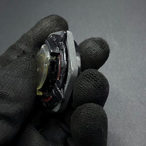K&ouml;nnte beinhalten: Ein schwarzes und silbernes Metall-Kreisel mit roten Akzenten. Der Kreisel wird in einer behandschuhten Hand gehalten.