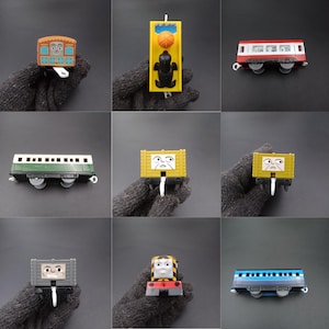 May include: A selection of toy train carriages, each with a distinct design and character face. The carriages include passenger and freight cars, plus a signal. The toys are made of plastic and are designed for children.