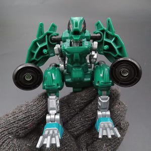 May include: A green and silver toy robot with black wheels and blue feet. The robot has a reptilian-like head and articulated limbs. The toy is made of plastic and appears to be a transforming figure.