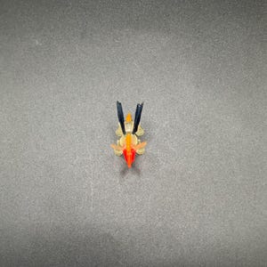 Puede incluir: Pequeña escultura abstracta y colorida. Presenta un diseño rojo, naranja y amarillo con detalles en negro. El objeto está sobre un fondo gris oscuro.