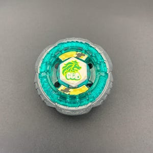 Könnte beinhalten: Ein kreisförmiges Kreiselspielzeug mit einem äußeren Ring aus silbernem Metall, einem mittleren Ring aus durchscheinendem Türkis und einem weißen sechseckigen Emblem in der Mitte. Das Emblem zeigt einen grünen Löwenkopf über dem Text GEO auf gelbem Tarnmuster.