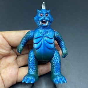 May include: A blue monster figurine with a textured, scaly appearance. The toy has red eyes, a white horn, and a detailed chest. The arms and legs are also textured, and the figure is held in a hand.