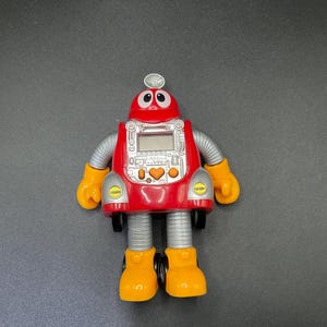 Puede incluir: Un robot de juguete rojo y amarillo con una pantalla digital en el pecho. El robot tiene brazos plateados, manos y pies amarillos y una pequeña antena en la parte superior. El diseño del robot incluye un corazón y detalles circulares.