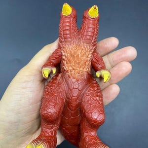 May include: A red and yellow monster toy with two heads, arms, and legs. The toy has a textured, scaly appearance with yellow claws and accents. The monster is a vintage collectible.