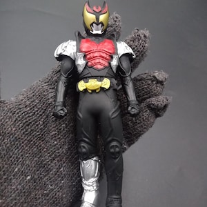 May include: An action figure of a character in a black and silver suit with a red chest plate and a gold helmet. The figure has a gold belt and silver boots. The figure is posed upright.