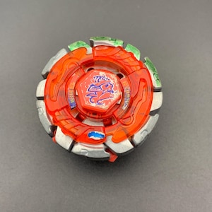 Puede incluir: Un juguete de peonza circular con un centro de plástico naranja translúcido y un emblema hexagonal central que muestra un gráfico de un dragón azul. El anillo exterior está construido con segmentos de metal plateado con detalles pintados en verde y azul.