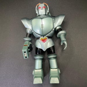 Puede incluir: Figura de acción de robot verde azulado metálico con un emblema en forma de corazón. El robot tiene una cabeza plateada con ojos rojos y una placa pectoral a juego. La figura tiene brazos y piernas articulados y un arma en el brazo derecho.