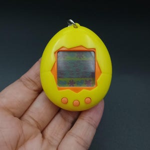 May include: A yellow and orange handheld digital pet toy. The egg-shaped device features a small screen displaying a pixelated image and three orange buttons below. A silver loop is at the top for attaching a keychain.