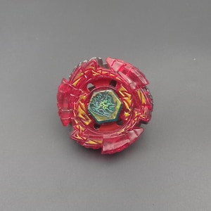 Könnte beinhalten: Ein kreisförmiges, rot-goldenes Kreiselspielzeug mit einem äußeren Ring aus durchscheinendem rotem Kunststoff mit gezackten Kanten und einem zentralen sechseckigen Emblem, das eine grün-goldene Drachengrafik auf dunklem Grund zeigt.