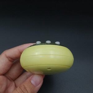 May include: A pale yellow, rounded electronic device with three small, gray buttons on top. The device is held in a hand against a dark background. The device has a small hole on the bottom.