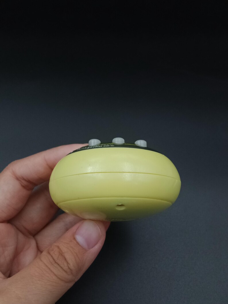 May include: A pale yellow, rounded electronic device with three small, gray buttons on top. The device is held in a hand against a dark background. The device has a small hole on the bottom.