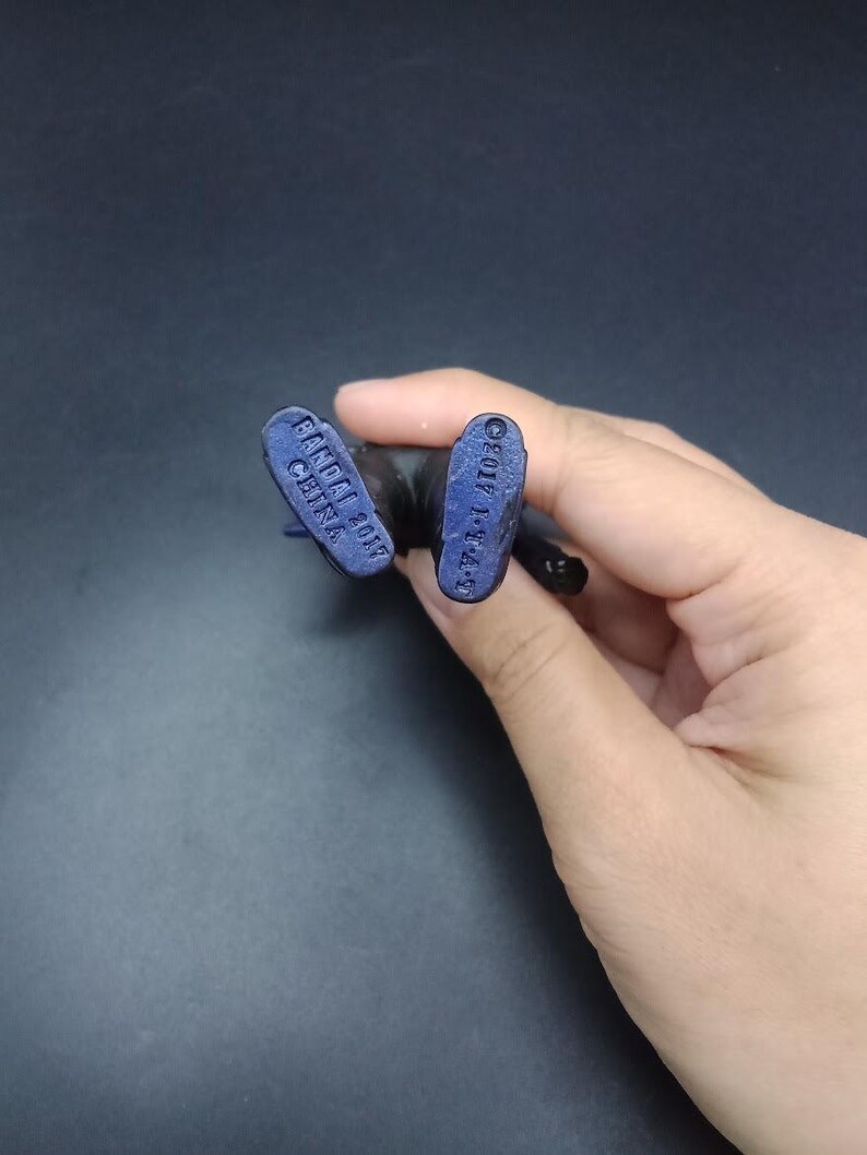 May include: Close-up of the feet of a small, dark blue action figure. The soles of the feet are visible, with the text "BANDAI 2017 CHINA" and "&copy;2017 T-T-A" imprinted on them. The figure is held against a dark background.