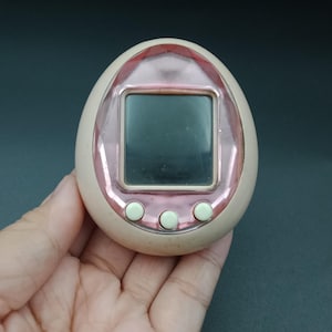 Könnte beinhalten: Ein Vintage-Handheld-Digital-Haustierspiel in einem hellrosa, eiförmigen Gehäuse mit einem metallisch rosa Rahmen um den Bildschirm. Es hat drei weiße Knöpfe. Das Gerät wird in einer Hand vor dunklem Hintergrund gehalten.