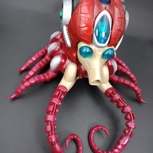 May include: A red and silver toy octopus with a helmet-like head and multiple tentacles. The toy has blue and green accents, with a beige face and a detailed, mechanical design. The tentacles are a darker shade of red and curl at the ends.