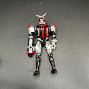 Könnte beinhalten: Metall-Roboter-Actionfigur mit rotem und schwarzem Torso, silbernen Armen und Beinen sowie einer weißen antennenartigen Struktur auf dem Kopf. Die Figur hat einen pistolenartigen Aufsatz am linken Arm und steht.