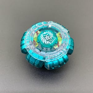 Puede incluir: Un juguete de peonza circular con una base de metal color verde azulado y un anillo exterior de plástico transparente. El perno central muestra un gráfico de cabeza de león blanco y las letras GT. Se ven pegatinas de camuflaje bajo el anillo.