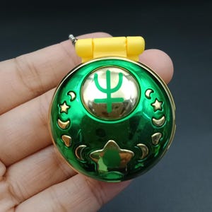 Puede incluir: Un compacto verde y dorado con una tapa amarilla. El compacto presenta un diseño de estrellas y lunas crecientes doradas en los bordes. Un símbolo dorado está en el centro.