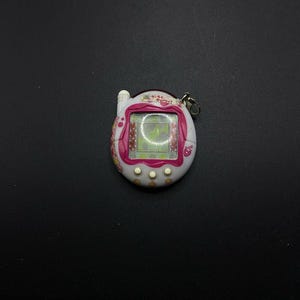 Puede incluir: Mascota digital blanca y rosa con una pantalla que muestra un corazón verde. La mascota tiene un botón rosa en la parte superior y tres botones blancos en la parte inferior. El texto en la mascota dice "Tamagotchi!"