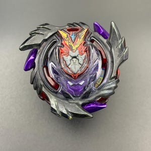 May include: A spinning top toy featuring a complex design with a metallic silver outer ring, purple accents, and a central red emblem with a V-shaped motif. The structure includes layered plastic components with sharp, angular edges and a dark colour palette.