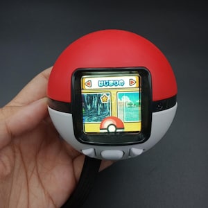 May include: A red, white, and black handheld electronic device resembling a Poké Ball. The device has a small screen displaying a colourful graphic. Three buttons are located below the screen. A black strap is attached to the device.