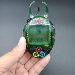 Puede incluir: Un juego electrónico portátil verde con un diseño de escarabajo. El dispositivo presenta una pequeña pantalla, botones coloridos y texto japonés. La parte superior tiene una estructura similar a una antena de tres puntas.