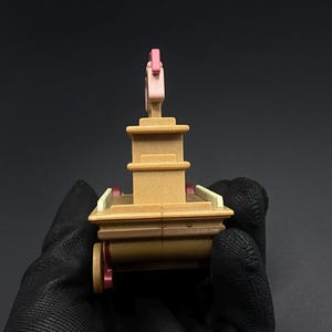May include: A miniature toy carriage in shades of brown, pink, and red. The toy has a multi-tiered structure with a pink decorative element on top and small red wheels. The carriage is held by a gloved hand against a dark background.