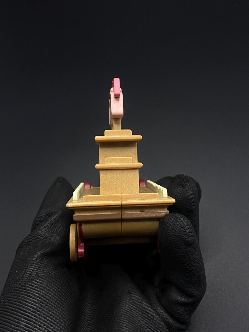 May include: A miniature toy carriage in shades of brown, pink, and red. The toy has a multi-tiered structure with a pink decorative element on top and small red wheels. The carriage is held by a gloved hand against a dark background.