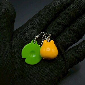 May include: A keychain featuring two charms: a bright green, circular charm with a cutout and a small, rounded orange charm. The charms are connected by a silver chain with small beads. The background is a dark, textured surface.