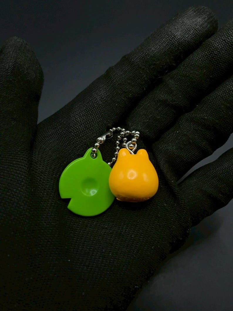 May include: A keychain featuring two charms: a bright green, circular charm with a cutout and a small, rounded orange charm. The charms are connected by a silver chain with small beads. The background is a dark, textured surface.