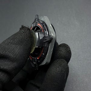 K&ouml;nnte beinhalten: Ein schwarzer und silberner Metall-Beyblade mit roten und orangen Akzenten. Der Beyblade wird in einer behandschuhten Hand gehalten.