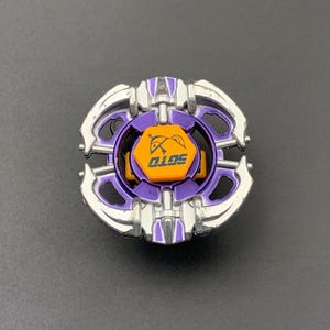 Puede incluir: Un juguete de peonza circular con un marco metálico morado y plateado y una tapa hexagonal naranja central. La tapa muestra un gráfico negro de un arco y una flecha sobre el texto SOTO impreso en una fuente negra, negrita y estilizada.
