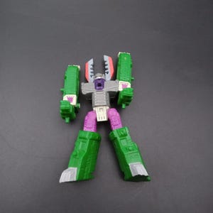 May include: A green, purple, gray, and white action figure of a robot. The robot has green legs and arms, a gray torso, and a purple head. The robot's arms are raised, and its legs are spread apart.