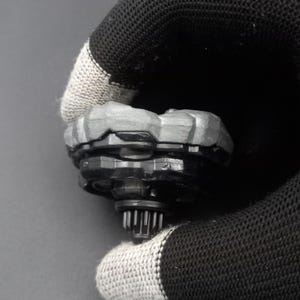 Pu&ograve; includere: Una trottola giocattolo nera e grigia con una parte superiore grigia strutturata e rocciosa e una base nera. Il giocattolo &egrave; tenuto da un guanto nero e grigio.