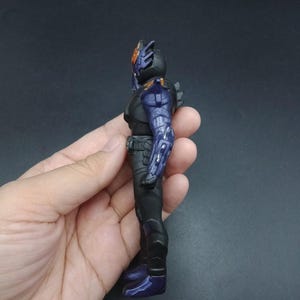 May include: A black and purple action figure with a detailed design. The figure has a helmet with orange accents, a black torso, and purple armor on the arms and legs. The figure is held in a hand against a dark background.