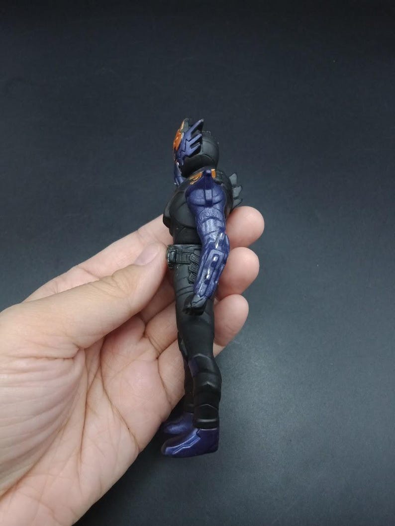 May include: A black and purple action figure with a detailed design. The figure has a helmet with orange accents, a black torso, and purple armor on the arms and legs. The figure is held in a hand against a dark background.