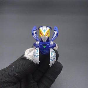 以下が含まれることがあります： 青と銀色のロボットのおもちゃ。黄色と白のアクセントがあり、翼と詳細なデザインが特徴です。小さな緑色のボタンが付いています。このロボットはコレクターズアイテムです。