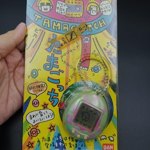 Puede incluir: Un juguete virtual Tamagotchi vintage en su embalaje original. El embalaje es amarillo con ilustraciones coloridas y texto japonés. El dispositivo Tamagotchi es verde y rosa con una pequeña pantalla. La marca es visible.