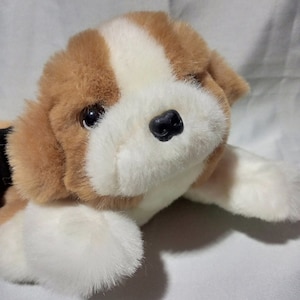 Op de afbeelding: Een bruine en witte pluche speelgoedhond met zwarte ogen en een zwarte neus. De hond ligt op zijn zij met zijn hoofd rustend op zijn poten.