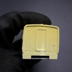 Puede incluir: Un pequeño camión de juguete amarillo con forma rectangular. La parte trasera del camión tiene una puerta y varias líneas horizontales. El camión está sobre un fondo oscuro.