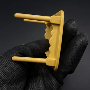 May include: A small, mustard-yellow shelf with two cylindrical pegs extending from the front. The shelf has a scalloped edge and is held by a gloved hand against a dark background. The shelf appears to be made of plastic.