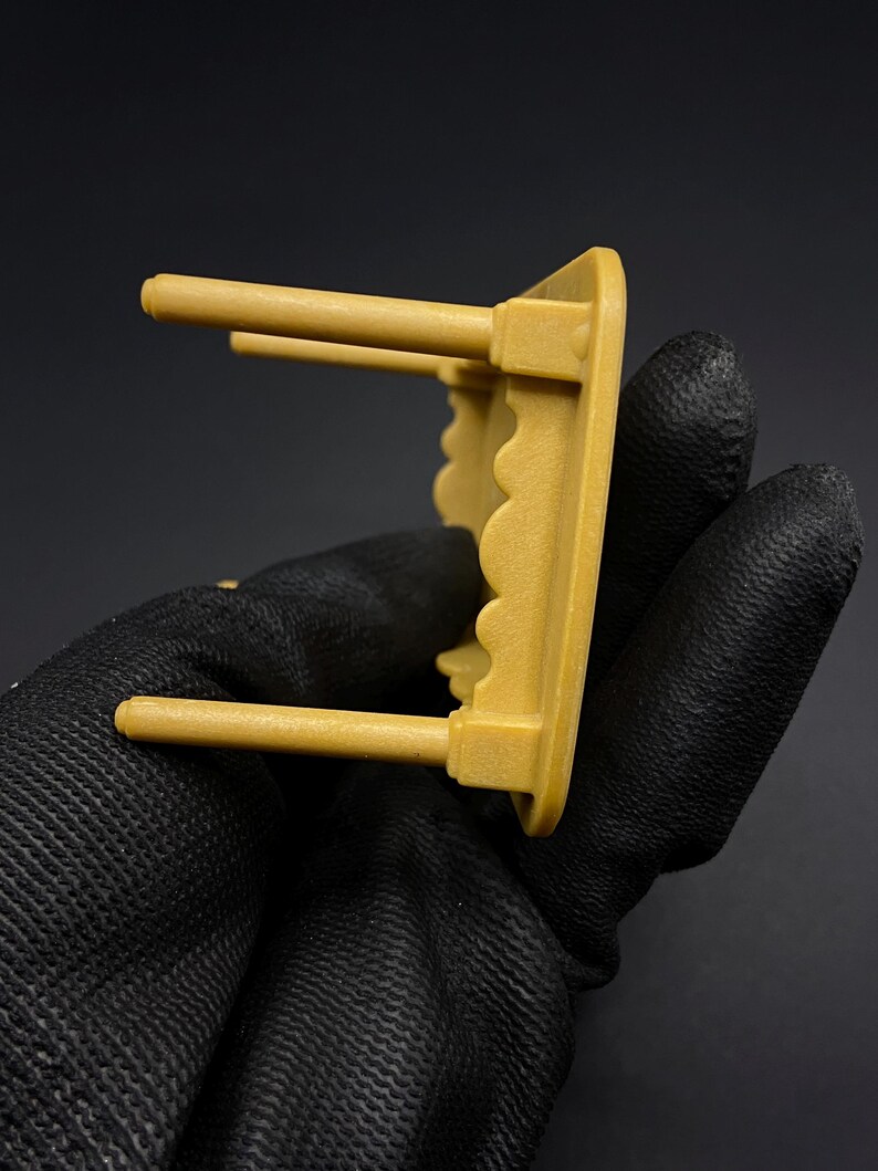 May include: A small, mustard-yellow shelf with two cylindrical pegs extending from the front. The shelf has a scalloped edge and is held by a gloved hand against a dark background. The shelf appears to be made of plastic.