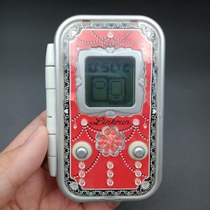 May include: A handheld electronic device with a red face and silver trim. The face features a digital display, two silver buttons, and the word "Linkrun". The device has a white side attachment and is held in a hand.