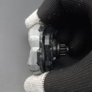 Pu&ograve; includere: Una trottola nera e grigia con un anello esterno grigio strutturato e un centro nero con un design a forma di ingranaggio. Il giocattolo &egrave; tenuto da una mano guantata.