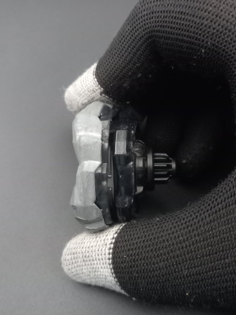 Pu&ograve; includere: Una trottola nera e grigia con un anello esterno grigio strutturato e un centro nero con un design a forma di ingranaggio. Il giocattolo &egrave; tenuto da una mano guantata.