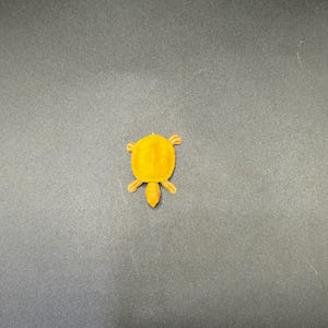 May include: A small, orange turtle figurine with a textured shell and four small legs. The turtle is made of a solid material and is set against a dark gray background. The figurine is a decorative item.