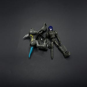 Peut inclure: Une figurine articulée grise et bleue démontée sur une surface gris foncé. La figurine a une tête bleue, des parties du corps grises et des accents bleus et jaunes. La figurine est en morceaux, avec les pièces éparpillées.