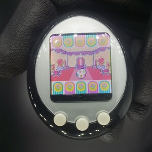 May include: A silver and black handheld electronic pet toy with a color screen displaying a cartoon image of a room with a throne, tables, and chairs. The screen has a pink, yellow, and blue border.