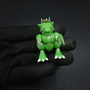 Puede incluir: Una figura verde articulada de una criatura reptiliana con detalles blancos en los pies. La figura tiene un accesorio de llavero y se encuentra sobre un fondo oscuro.
