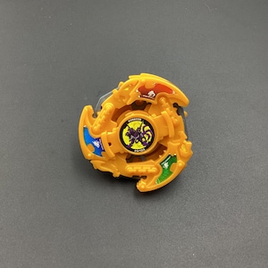May include: A yellow plastic Beyblade with a purple and black design in the center. The Beyblade has four blades, each with a different color: blue, red, green, and yellow.